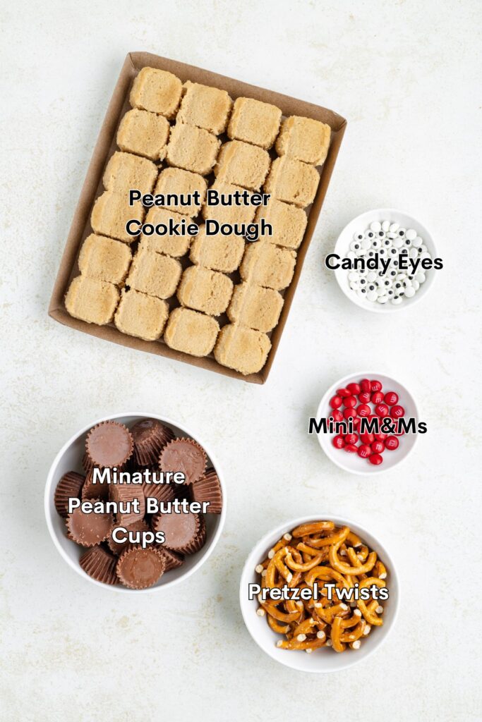 Top view of baking ingredients: a tray of peanut butter cookie dough squares, bowls of candy eyes, mini red M&Ms, miniature peanut butter cups, and pretzel twists, arranged on a white surface.