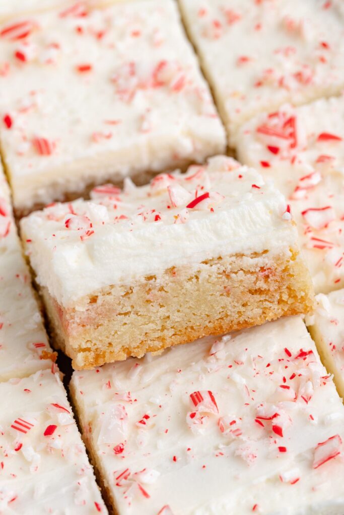 Close-up of frosted blondies topped with crushed peppermint candies. One blondie is slightly raised, showing a dense, moist texture beneath the white frosting and red candy pieces.