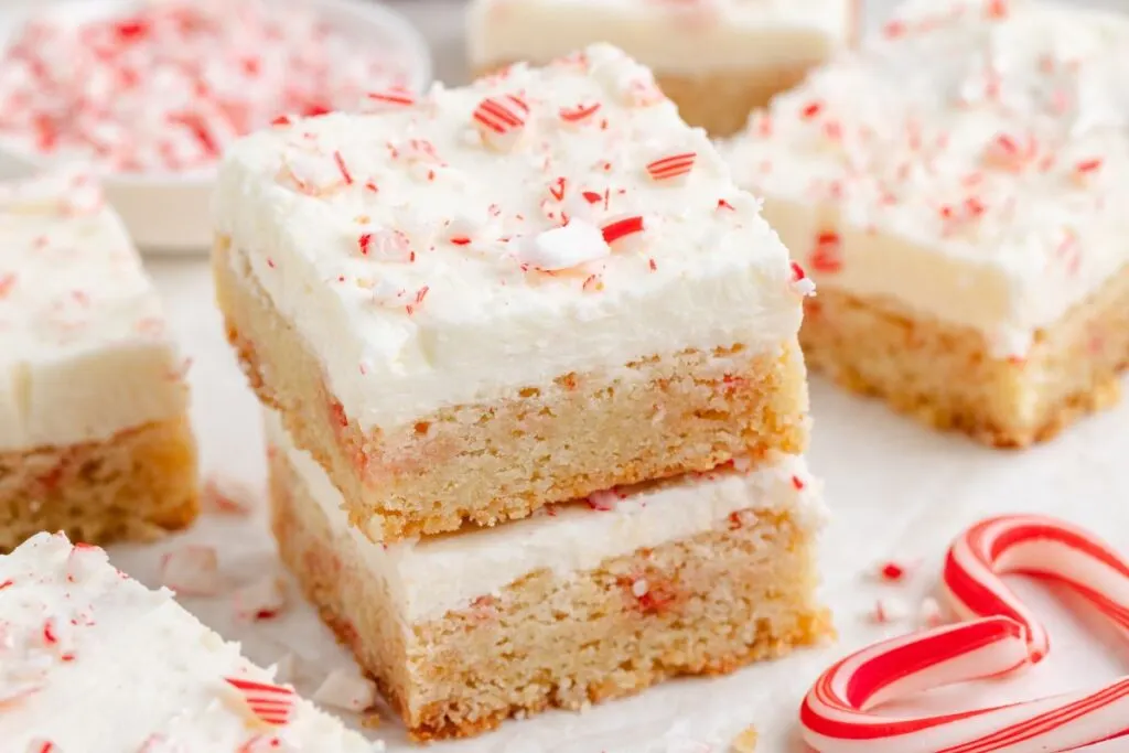 Two square peppermint sugar cookie bars stacked on top of each other, topped with creamy white frosting and crushed peppermint candies. A heart-shaped candy cane lies beside them, with more cookie bars and candy pieces in the background.