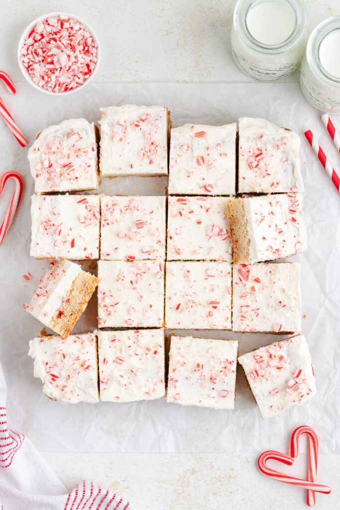 Squares of peppermint sugar cookies on parchment paper, topped with white frosting and crushed candy canes. Nearby are candy canes, a small bowl of peppermint pieces, and two jars of milk.