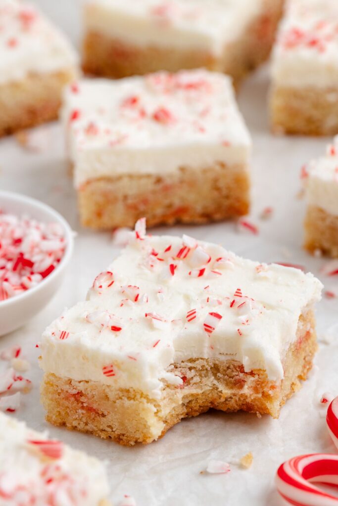 A close-up of peppermint sugar cookie bars with creamy white frosting and crushed peppermint pieces on top. One bar has a bite taken out, revealing a dense, moist texture. Other bars and peppermint candies are visible in the background.