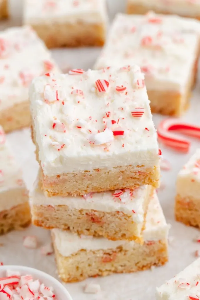 A stack of peppermint sugar cookie bars with white frosting and crushed candy cane pieces on top. More cookie bars and candy canes are spread across the background.