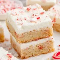 Two stacked peppermint sugar cookie bars with creamy white frosting and crushed peppermint candy on top. More bars and peppermint pieces are visible in the background.