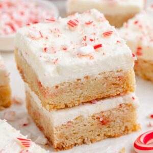 Two stacked peppermint sugar cookie bars with creamy white frosting and crushed peppermint candy on top. More bars and peppermint pieces are visible in the background.