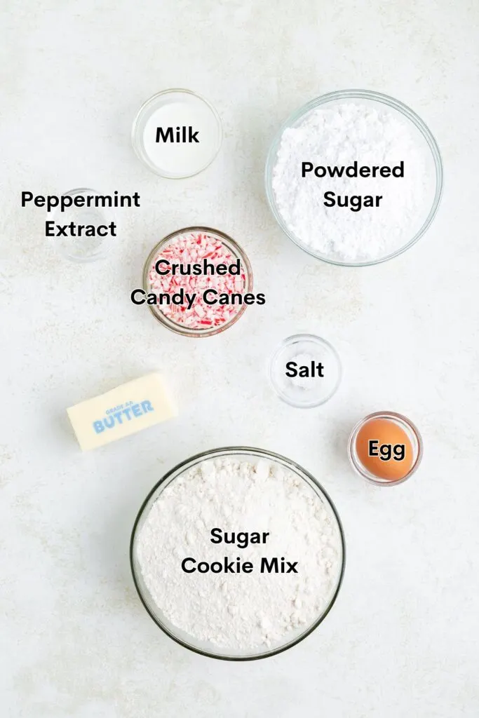 Top-down view of various baking ingredients arranged on a light surface. Includes bowls of powdered sugar, sugar cookie mix, peppermint extract, crushed candy canes, milk, salt, an egg, and a stick of butter. Each item is labeled clearly.