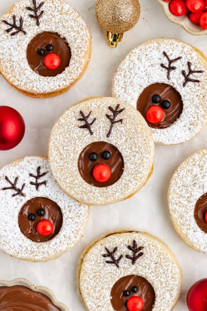 Festive reindeer linzer cookies decorated with nutella, red candy noses, and piped antlers. The cookies are dusted with powdered sugar and surrounded by red and gold Christmas ornaments on a light background.