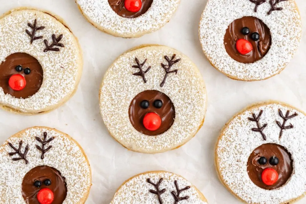 Seven festive linzer cookies decorated like reindeer with nutella centers, red candy noses, and black candy eyes. They are dusted with powdered sugar and arranged on a light surface.