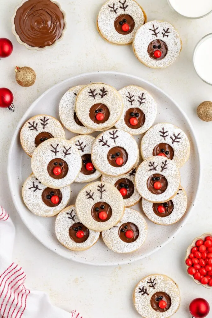 A plate of reindeer-themed cookies decorated with Nutella centers, red candy noses, and icing antlers. Surrounded by bowls of chocolate spread, glasses of milk, red ornaments, and festive decorations on a white surface.