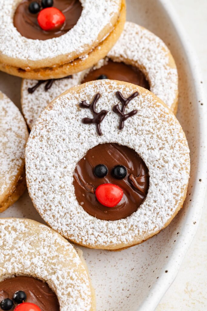 A plate of Nutella reindeer linzer cookies decorated with powdered sugar. Each cookie has a cut-out center filled with chocolate, featuring a red candy nose and chocolate eyes, resembling a reindeer. Chocolate antlers are piped above the eyes.