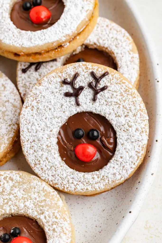 A plate of Nutella reindeer linzer cookies decorated with powdered sugar. Each cookie has a cut-out center filled with chocolate, featuring a red candy nose and chocolate eyes, resembling a reindeer. Chocolate antlers are piped above the eyes.