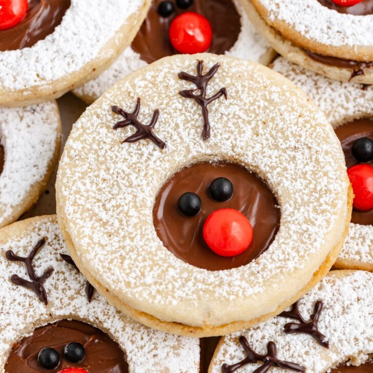 Festive cookies topped with powdered sugar, chocolate spread, and decorated to look like reindeer faces with chocolate antlers, red candy noses, and black candy eyes.