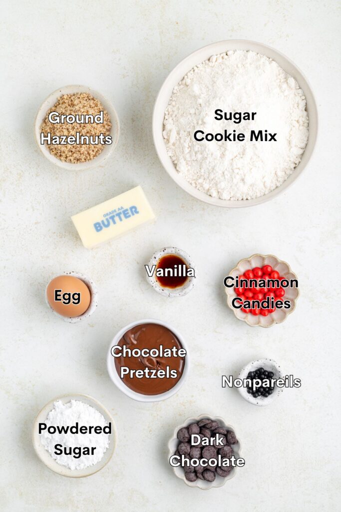 A flat lay of baking ingredients: sugar cookie mix, ground hazelnuts, a stick of butter, an egg, vanilla, chocolate pretzels, powdered sugar, dark chocolate, cinnamon candies, and nonpareils.