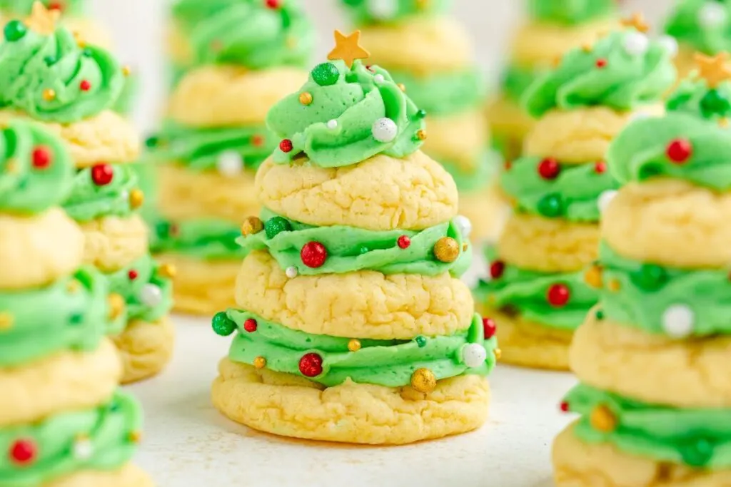 Stacked Christmas tree cookies feature layers of green frosting and round sprinkle decorations in red, white, and gold. Topped with a small gold star, these festive treats are artfully arranged on a light surface.