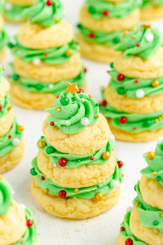 Festive cookies shaped like Christmas trees, stacked in layers with green frosting. Decorated with colorful edible beads and topped with a small star. Placed on a light surface.