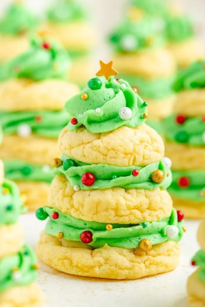 A festive stack of cookies shaped like a Christmas tree, layered with green frosting and decorated with colorful sprinkles and a small star on top. More cookie stacks are blurred in the background.