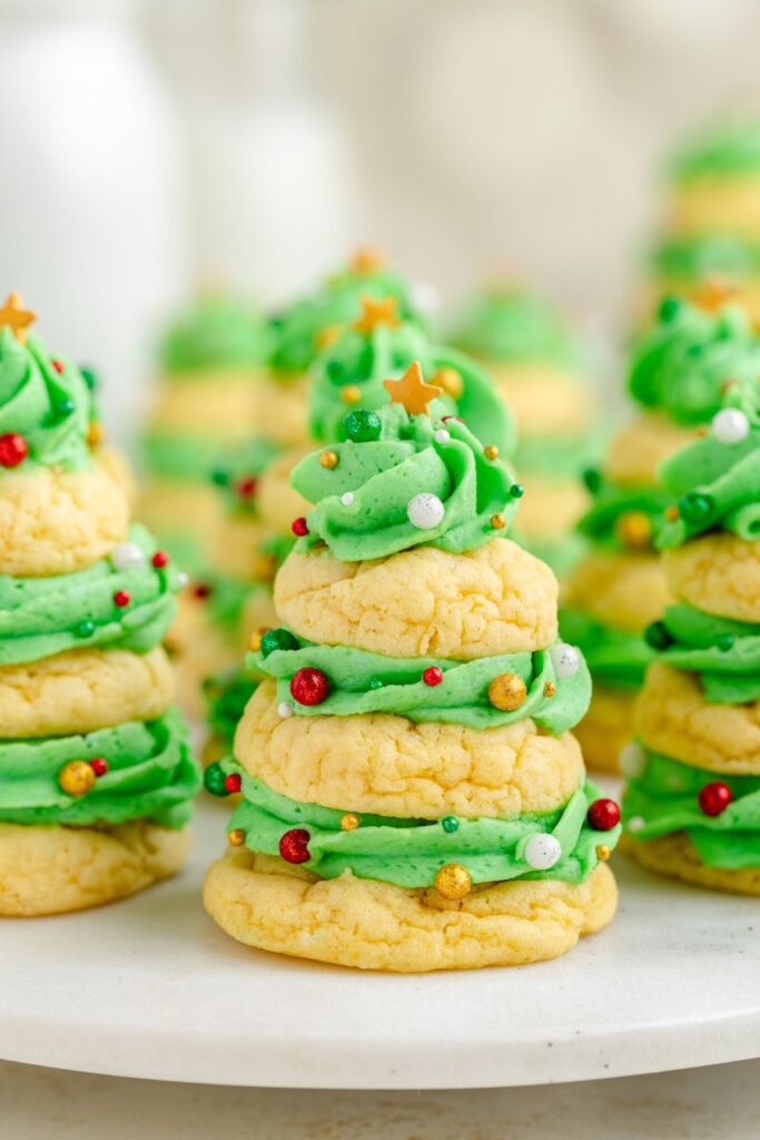 A festive dessert featuring cookies stacked like Christmas trees, with green frosting and decorated with red, white, and gold sprinkles, topped with a small gold star. They are displayed on a white surface.