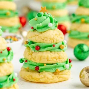 A festive cookie shaped like a Christmas tree, made from stacked yellow cookies with green frosting. It is decorated with colorful edible beads and topped with a small star. Other similar cookies and decorative ornaments are blurred in the background.