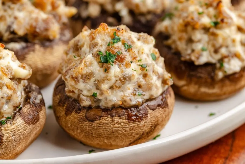 A close-up of three ingredient sausage stuffed mushrooms on a white plate. The mushrooms are filled with a mixture of sausage and cream cheese, garnished with chopped parsley, with the filling boasting a rich, golden-brown texture.