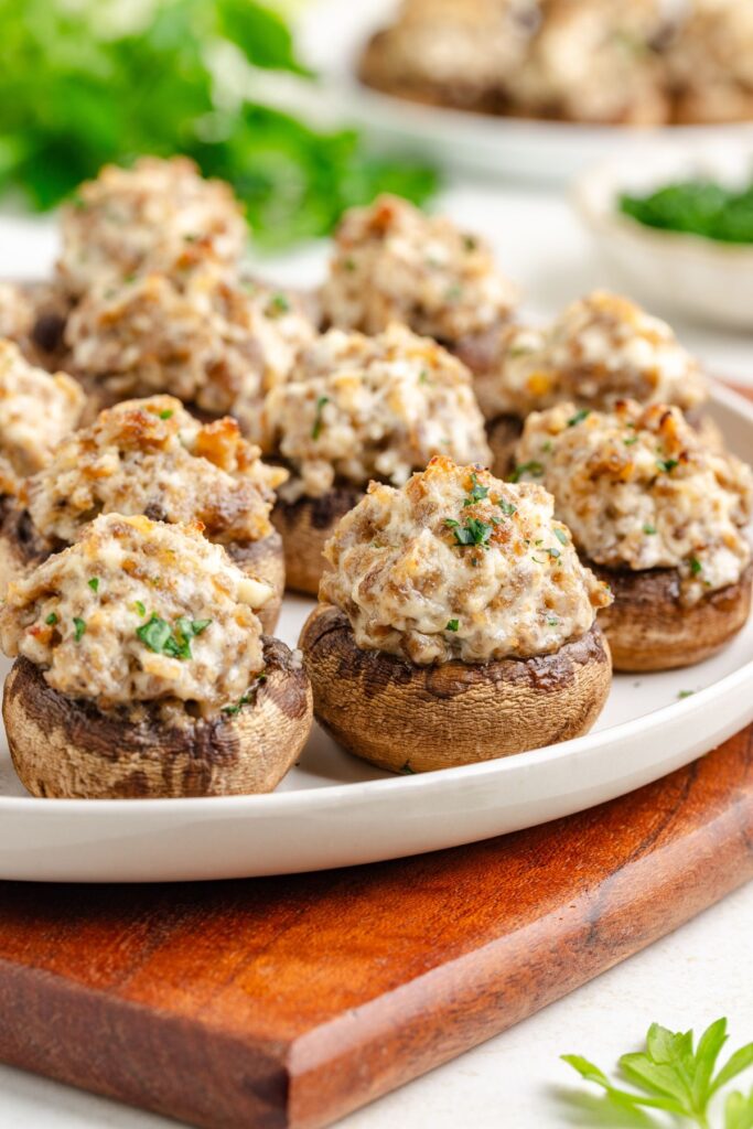 A plate of sausage stuffed mushrooms topped with a browned filling, placed on a wooden board. Fresh parsley is visible in the background, adding a hint of green to the scene.