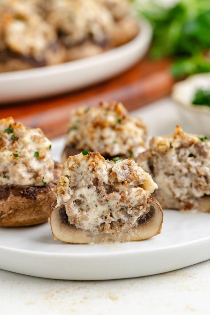 Close-up of stuffed mushrooms, with a creamy sausage filling and garnished with herbs, on a white plate. A plate of more stuffed mushrooms is blurred in the background.