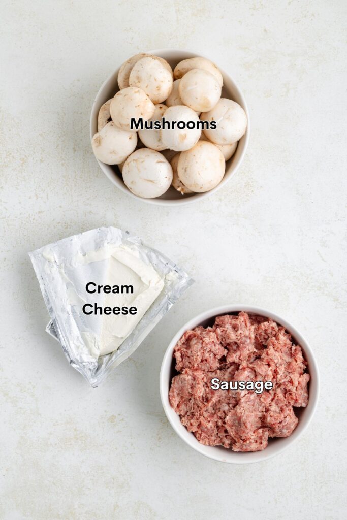 Three labeled items on a light surface: a bowl of whole mushrooms, a wedge of cream cheese, and a bowl of raw sausage meat.