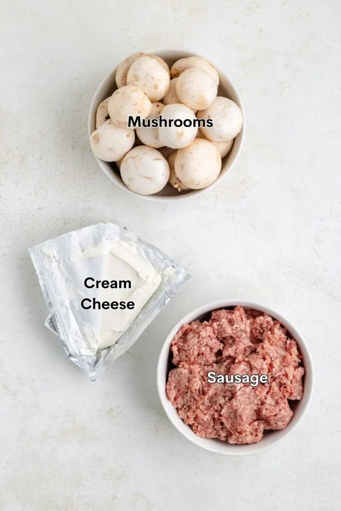 Three labeled items on a light surface: a bowl of whole mushrooms, a wedge of cream cheese, and a bowl of raw sausage meat.