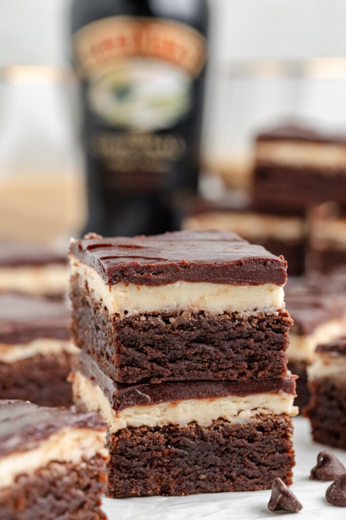 A stack of layered Baileys Brownies with a bottle of Irish cream liqueur in the background. They feature a creamy Irish cream layer sandwiched between rich chocolate, topped with a glossy chocolate ganache.