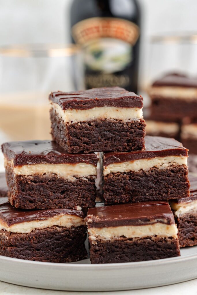 A plate of Baileys brownies with creamy layers on top. The brownies are stacked in three tiers. In the blurred background, theres a Baileys Irish Cream bottle and glasses, suggesting a casual dining setup.