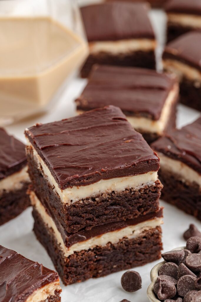 Close-up of Baileys Irish Cream brownies with layers of creamy filling and topped with a glossy chocolate glaze. Some brownies are stacked, while chocolate chips are scattered nearby.