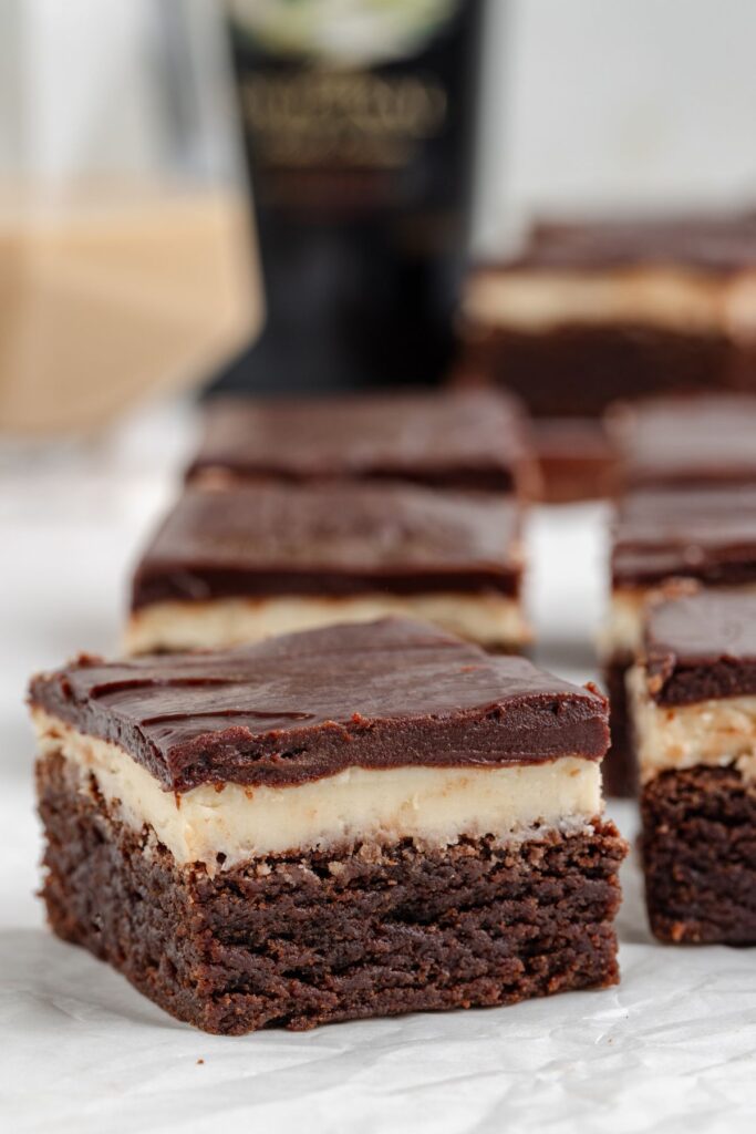 Close-up of Baileys brownies on a white surface, boasting a chocolate base, creamy middle infused with Baileys, and rich chocolate ganache on top. The background features a blurred bottle of Baileys liquor.