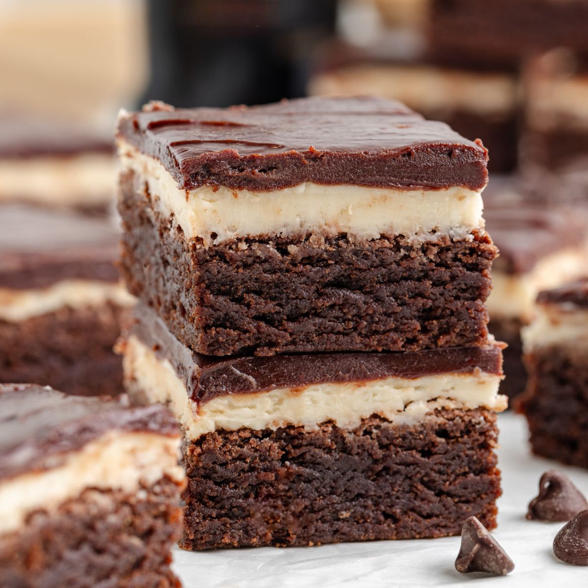 A stack of two Baileys Brownies sits on a marble surface. Each square boasts a rich brownie base, a creamy Baileys-infused middle layer, and a glossy chocolate top. Chocolate chips are scattered around, while other dessert pieces blur into the background.