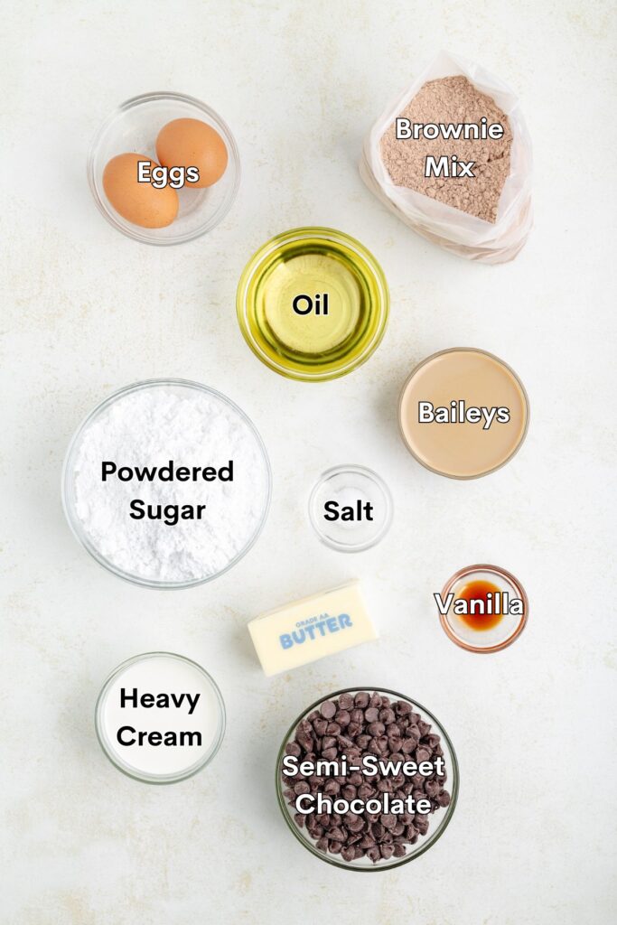 Top view of baking ingredients on a light surface: eggs, brownie mix, oil, Baileys, powdered sugar, salt, vanilla extract, heavy cream, unsalted butter, and semi-sweet chocolate chips.