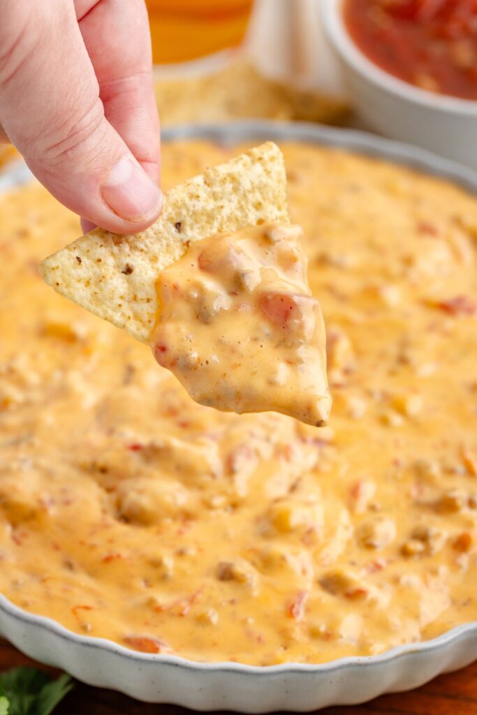 A hand holds a tortilla chip dipped in creamy Crockpot Rotel Sausage Dip, speckled with visible chunks of tomato and peppers.