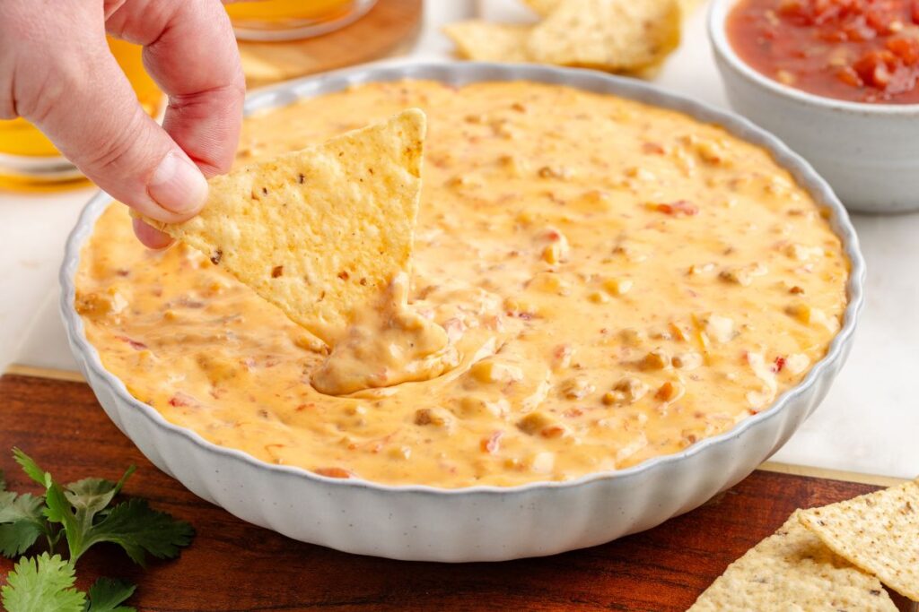 A hand dips a tortilla chip into a bowl of cheesy sausage dip, with flecks of tomato visible. A small bowl of red salsa and cilantro leaves are in the background on a wooden surface.