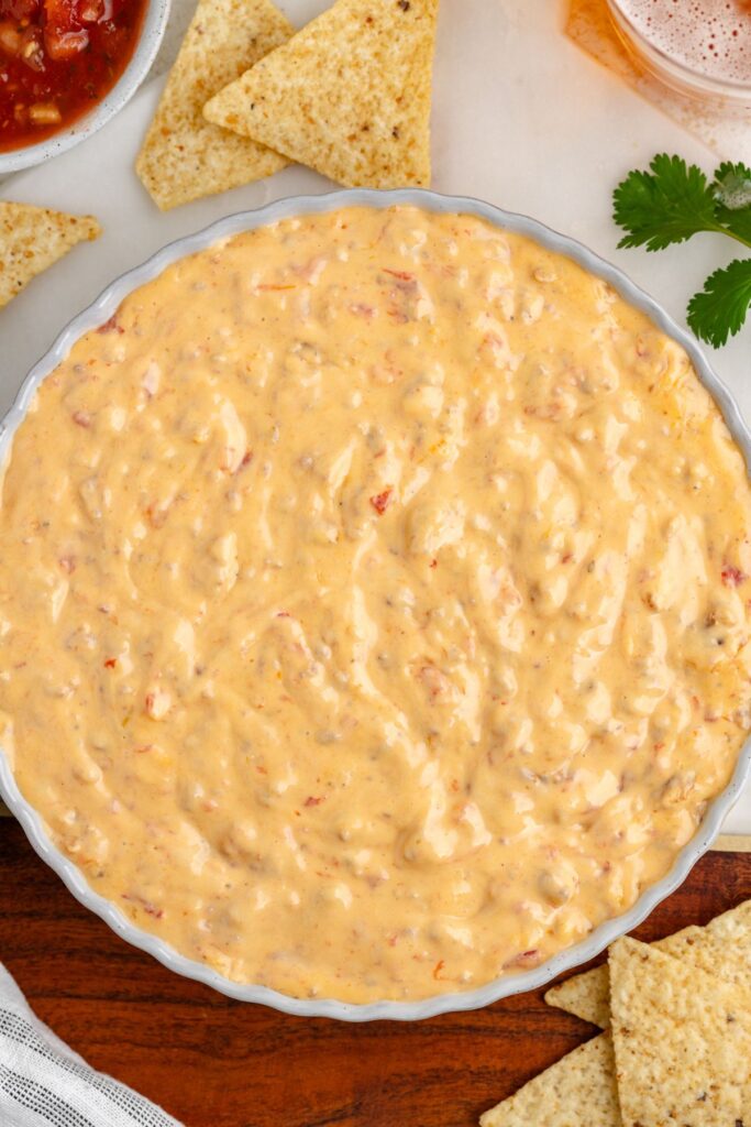 A dish filled with Velveeta Sausage Dip is surrounded by tortilla chips. Theres a small bunch of cilantro and a glass with a drink in the background.