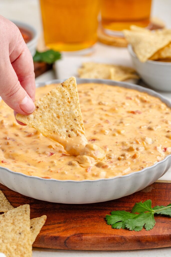 A hand dips a tortilla chip into a bowl of Crockpot Rotel sausage dip, with visible chunks of sausage and tomatoes. The bowl rests on a wooden board with a cilantro leaf nearby.