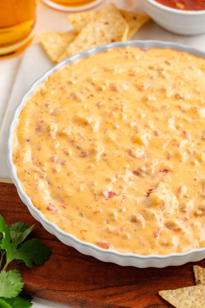 A bowl of sausage cheese dip with visible chunks of tomato and peppers sits on a wooden board. Tortilla chips and a small bowl of salsa are nearby, with a sprig of cilantro in the foreground. Glasses of beer are partially visible in the background.