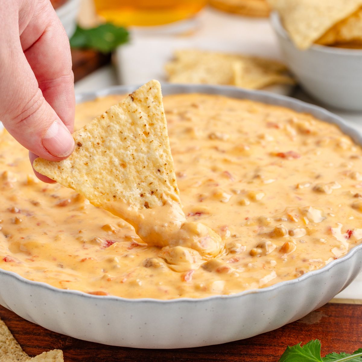 A hand dips a tortilla chip into a bowl of creamy slow cooker Sausage Dip, speckled with bits of tomato and peppers. Additional tortilla chips surround the bowl in the softly blurred background.