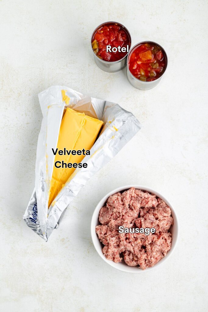 Top-down view of ingredients for a dip on a white surface: a partially unwrapped block of Velveeta cheese, a bowl of ground sausage, and two small tins of Rotel diced tomatoes and green chilies.