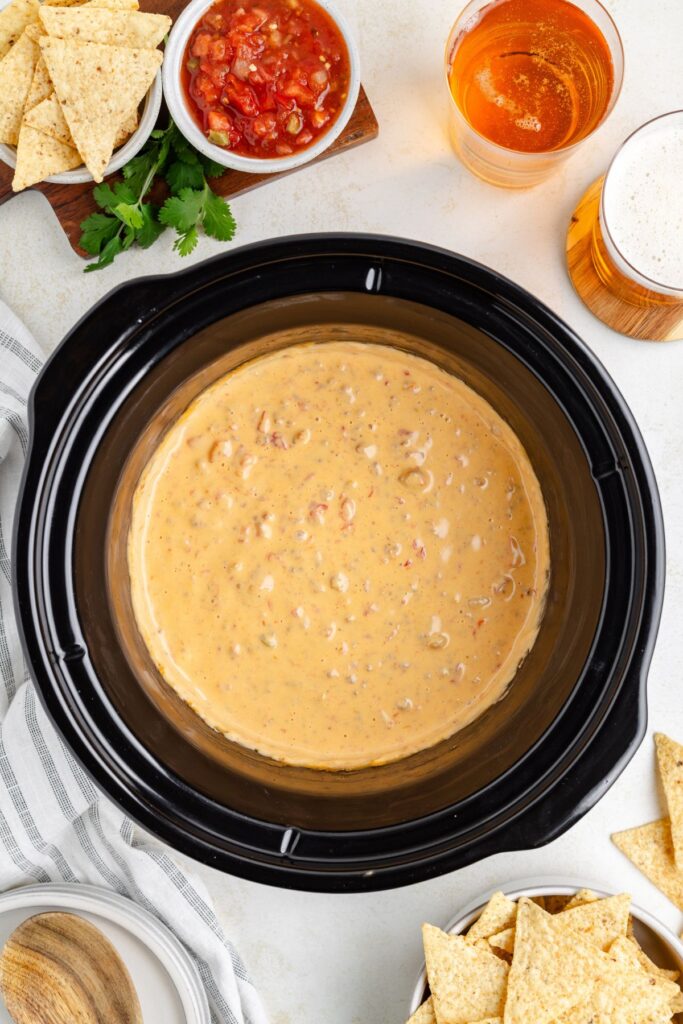 Slow cooker filled with creamy cheese dip, surrounded by tortilla chips, a bowl of salsa, a glass of beer, and a bunch of cilantro on a white surface. A striped cloth napkin and a wooden spoon are also visible.