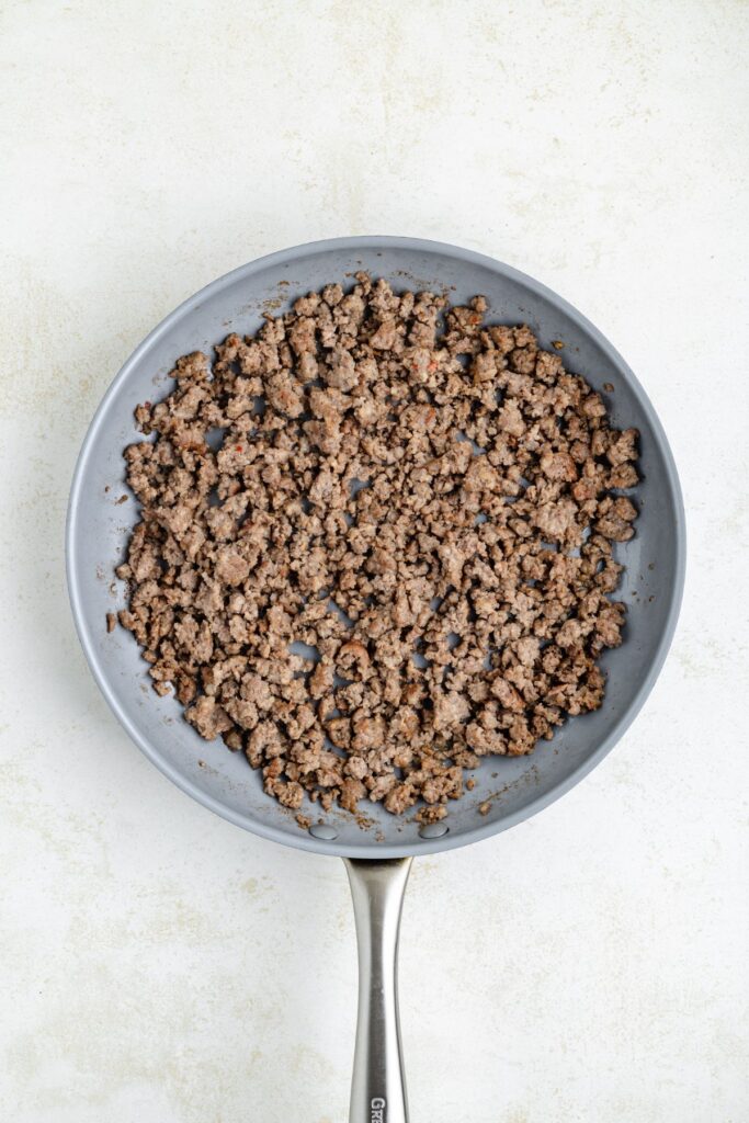 A frying pan filled with browned ground sausage on a light textured surface. The meat is evenly spread and cooked until slightly crispy. The pan has a silver handle.