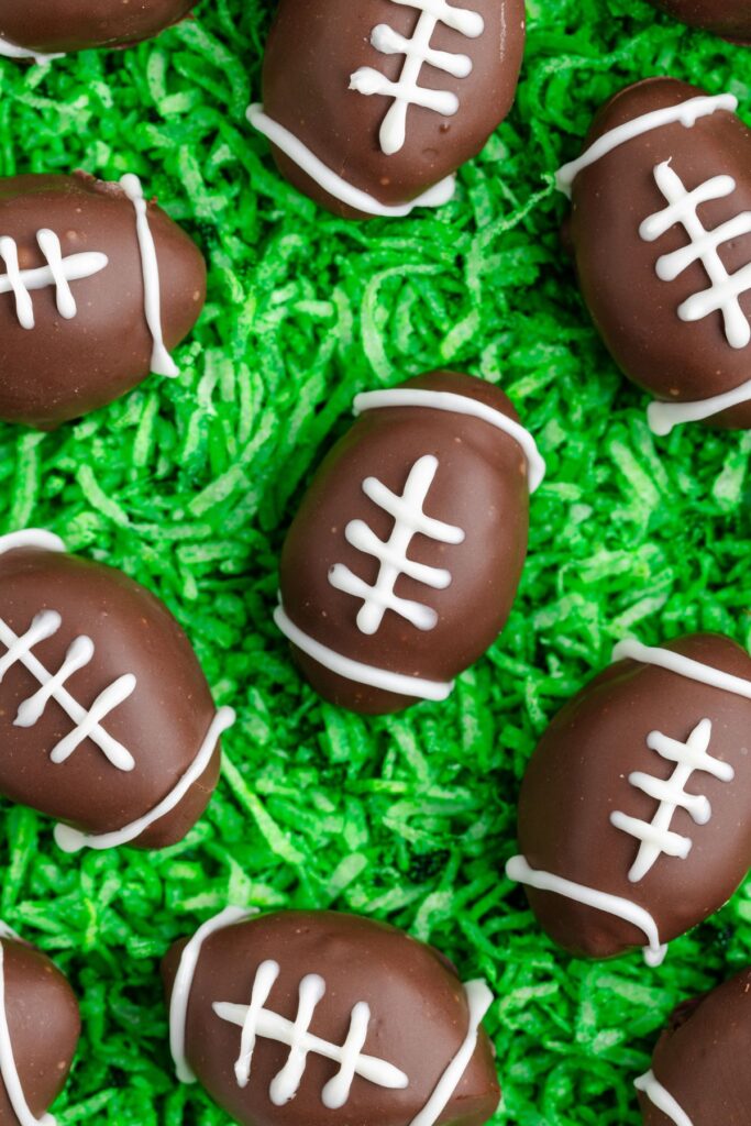 Chocolate-covered Oreo Truffles like footballs with white chocolate laces are placed on a bed of green shredded coconut, resembling grass.