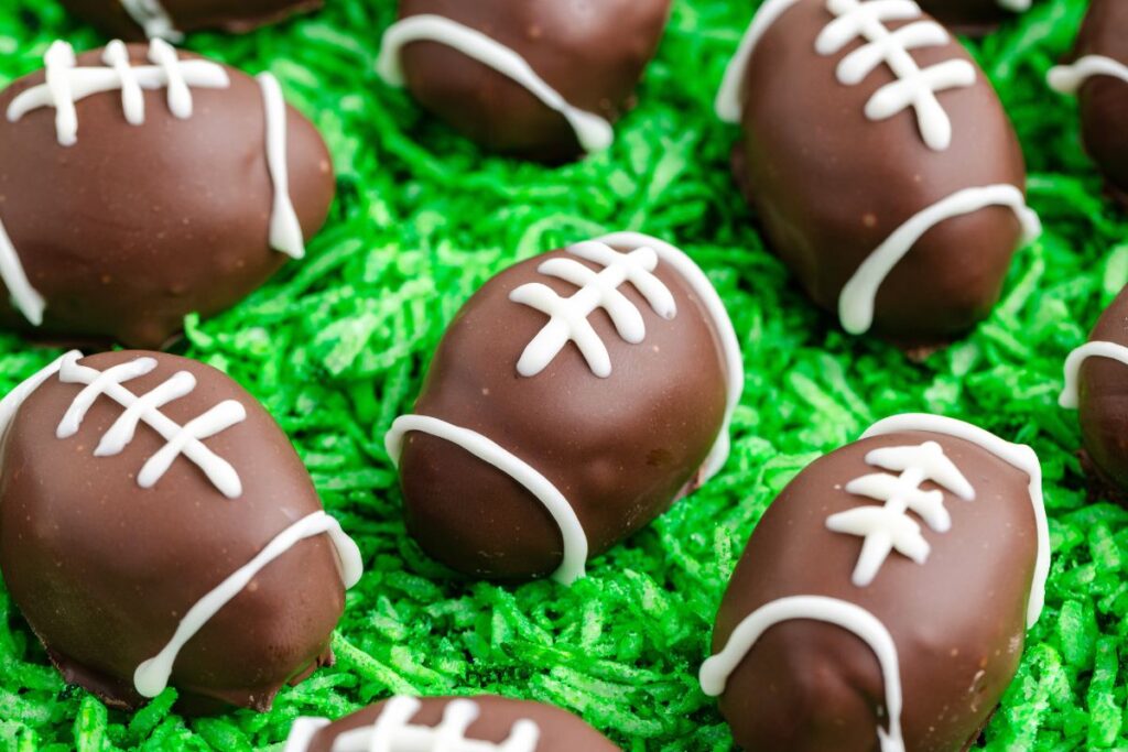 Chocolate treats shaped like footballs, decorated with white piping to resemble laces, are arranged on a bed of green shredded coconut, mimicking grass.