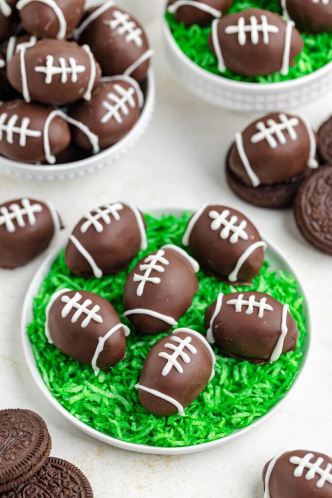 Chocolate Oreo Truffles shaped like footballs with white laces are displayed on a plate filled with green shredded coconut, resembling grass. Additional treats and sandwich cookies are in the background.