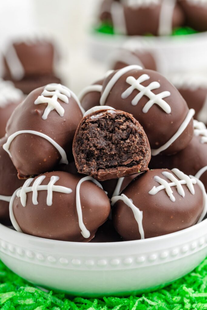 A white bowl filled with chocolate truffles decorated to look like footballs using white icing. One truffle is cut in half, revealing a dark, rich interior. The bowl is placed on a bed of green shredded material, resembling grass.