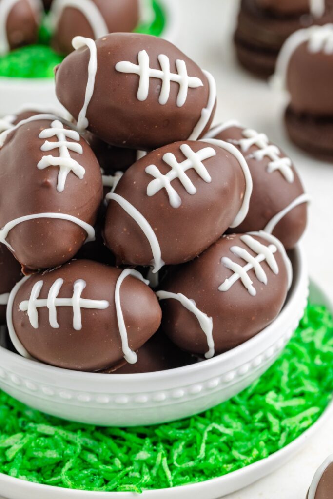 A bowl of chocolate candies shaped like footballs with white icing laces, placed on green shredded coconut resembling grass. More candies are visible in the background.