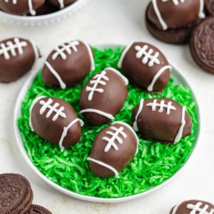 A plate of chocolate-covered Oreo truffles shaped like footballs, is arranged on shredded green coconut resembling grass. Additional treats and cookies are visible in the background.