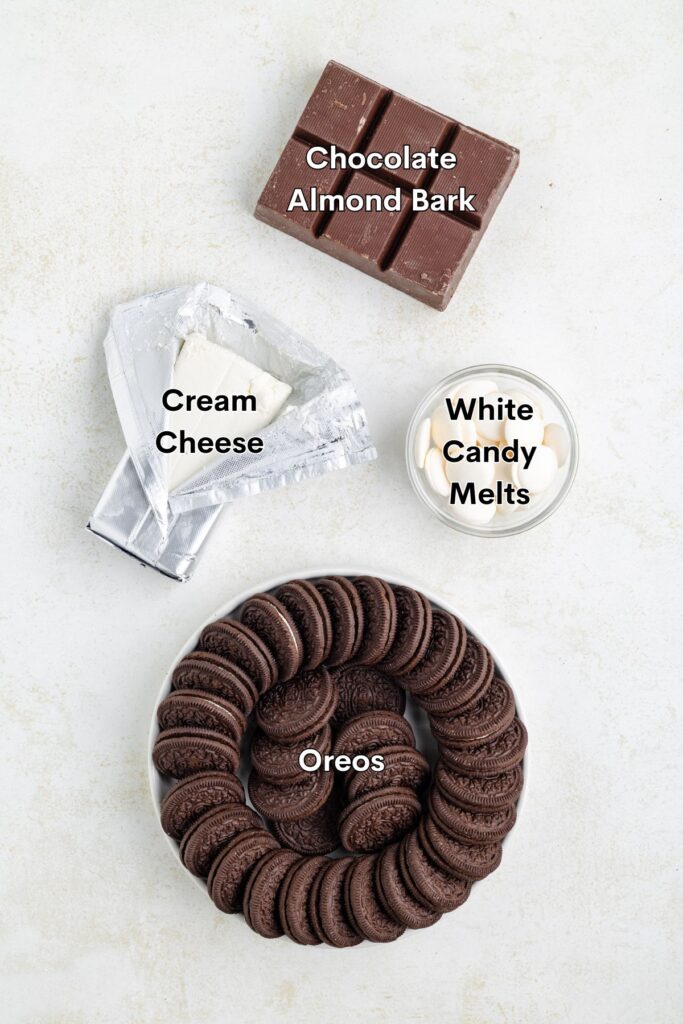 A top-down view of ingredients for a dessert. Includes a block of chocolate almond bark, a partially unwrapped block of cream cheese, a bowl of white candy melts, and a round plate of Oreos arranged in a circle.