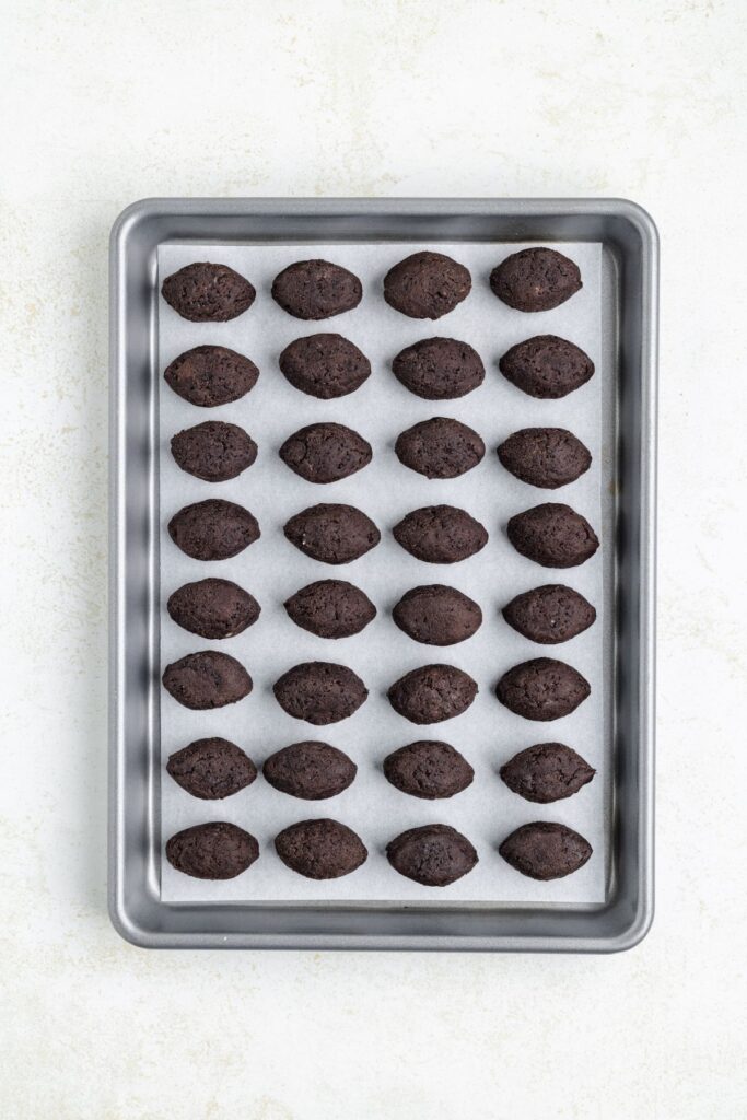 A baking tray holds five rows of football-shaped, Oreo truffle dough arranged neatly on parchment paper. The background is a light, textured surface.