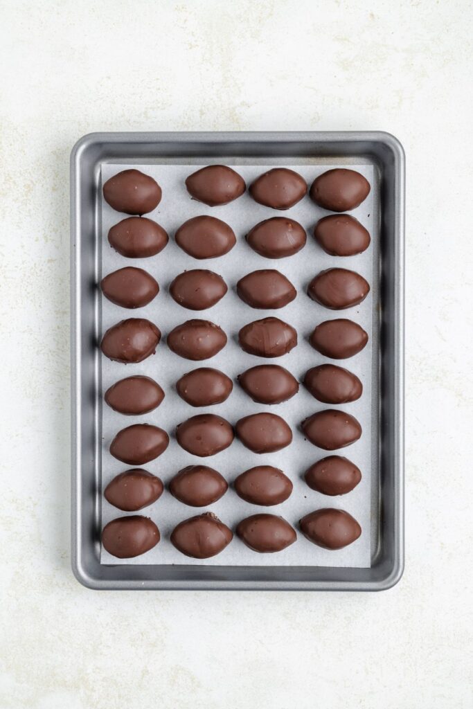 A baking tray lined with parchment paper holds five rows of chocolate-covered, football-shaped truffles arranged neatly. The tray is placed on a light-colored surface.